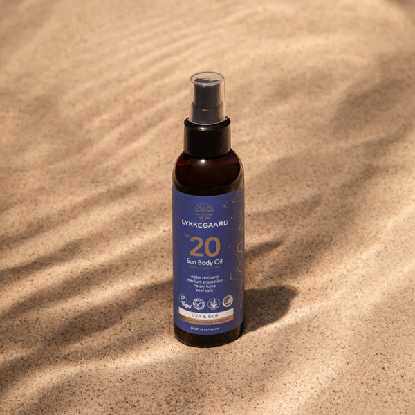 LYKKEGAARD Sun Body Oil SPF 20 bottle on sandy beach, offering lightweight sun protection and a radiant glow.