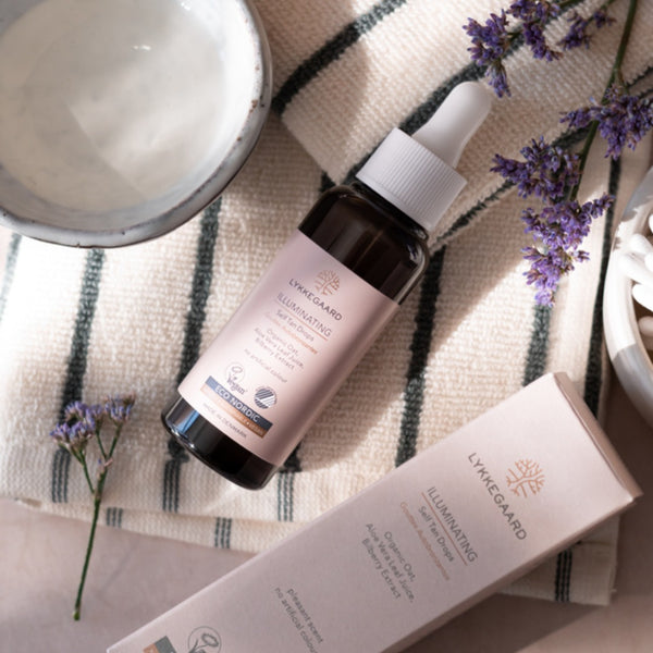 LYKKEGAARD Illuminating Self Tan Drops bottle with packaging and lavender on a textured cloth.